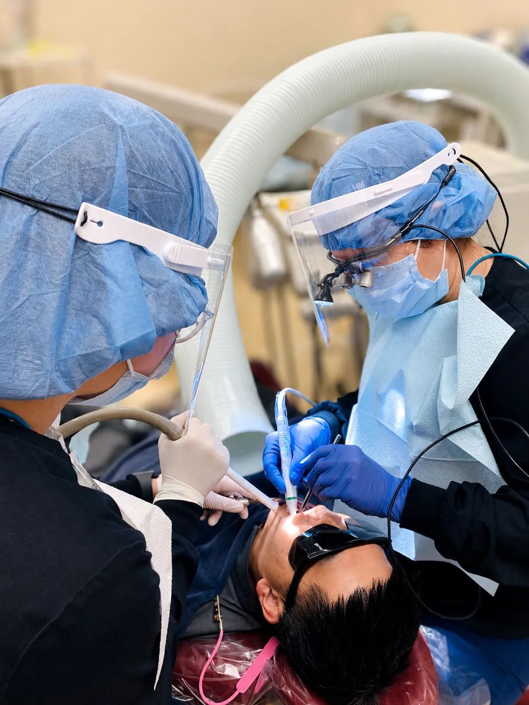 Two SoftDental team members in full PPE treating a patient — one administering anesthesia, the other holding the suction